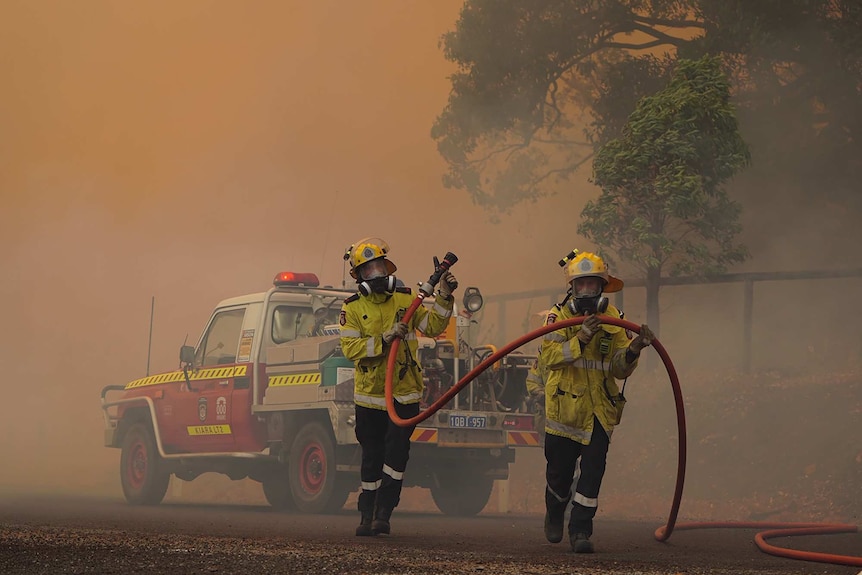 Two firefighters carry a fire hose away from a four-wheel drive with thick smoke surrounding them from a bushfire.