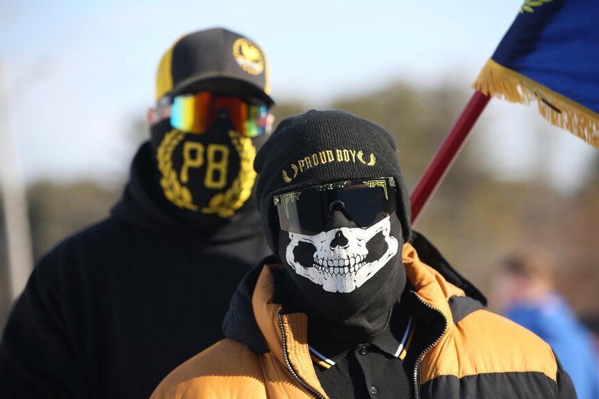 Two people are seen outdoors wearing masks and black and gold Proud Boys insignia