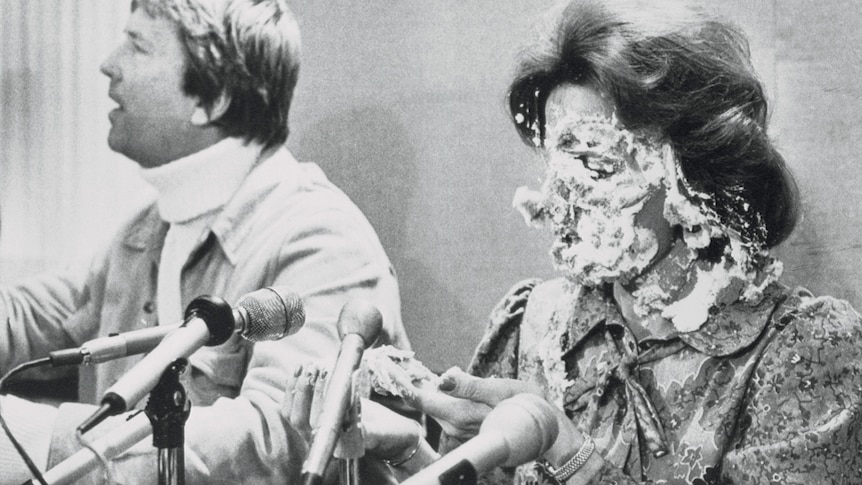 A 1970s black and white photo of a woman at a news conference, her face covered in pie, as a man sits next to her