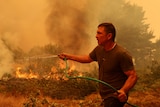 A man holds a hose. A fire burns behind him. 