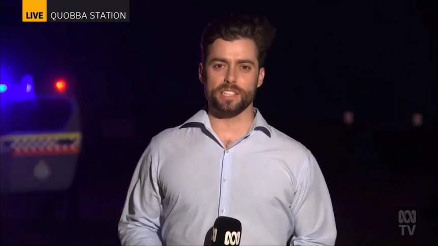 A journalist with an ABC microphone talks to camera at night with police lights behind him.