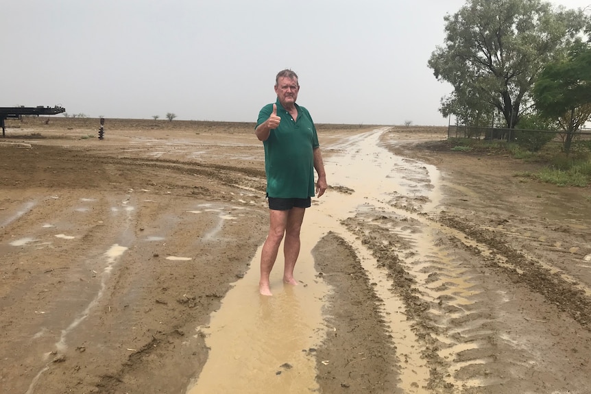 Man in shorts and a green shirt stands barefoot in soaked ground pointing thumbs up at camera