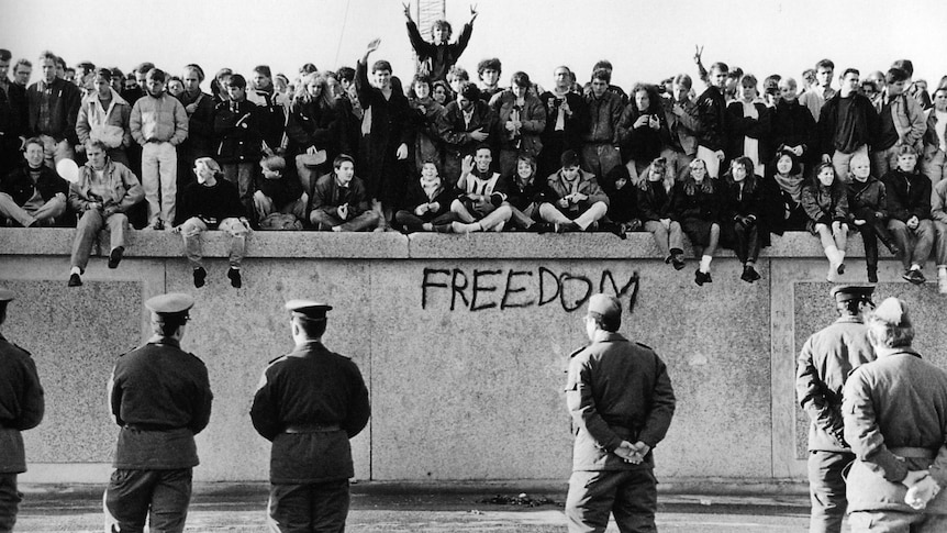 East German students sit atop the Berlin Wall in front of border guards