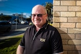 A man smiles and leans against a brick wall