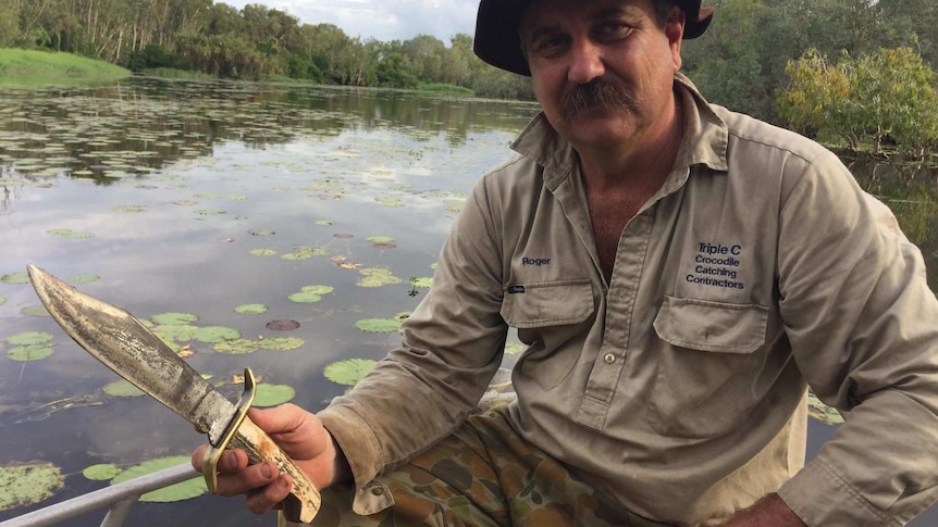 Roger Matthews sit in his boat on a billabong holding a large Bowie knife