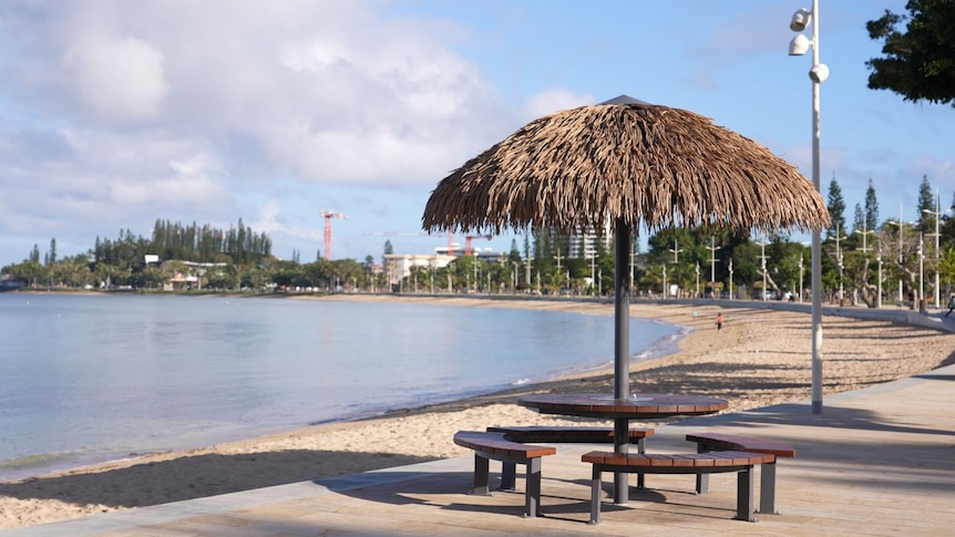 An empty table under a thatched umbrella with an empty beach in the background.
