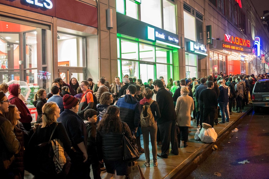 A queue of Star Wars fans lining up along a pavement at night outside a cinema in New York City.
