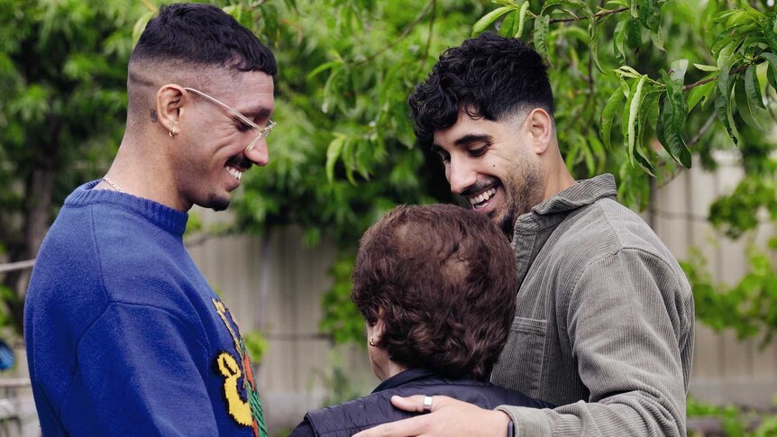 Two brothers smile down at a shorter, older woman. The three are hugging in a garden.