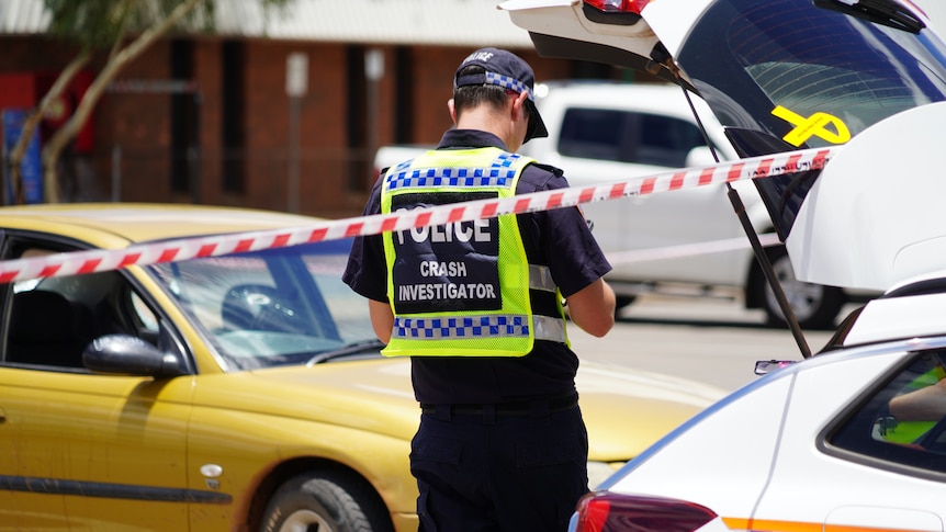 A police officer standing between a police vehicle and another gold car in a car park, with crime tape in the foreground.