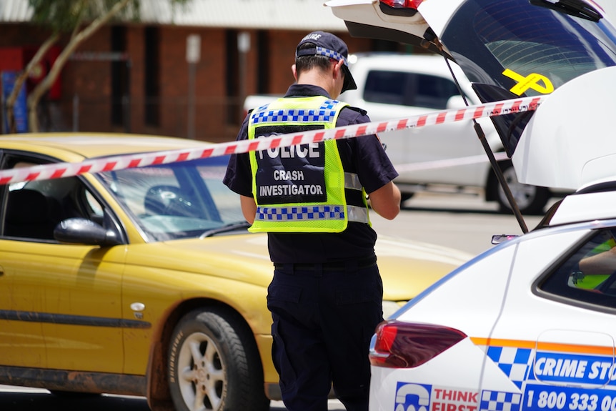 A police officer standing between a police vehicle and another gold car in a car park, with crime tape in the foreground.