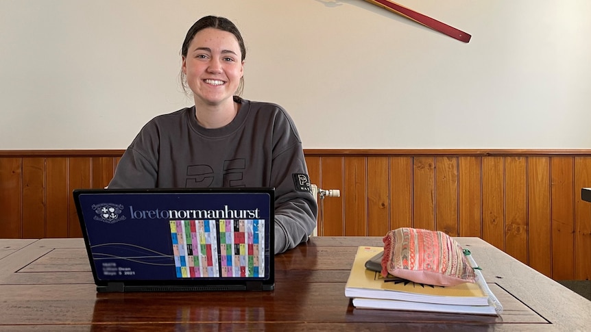 A brownhaired girl smiles at the camera sitting behind a laptop with notepads on a desk