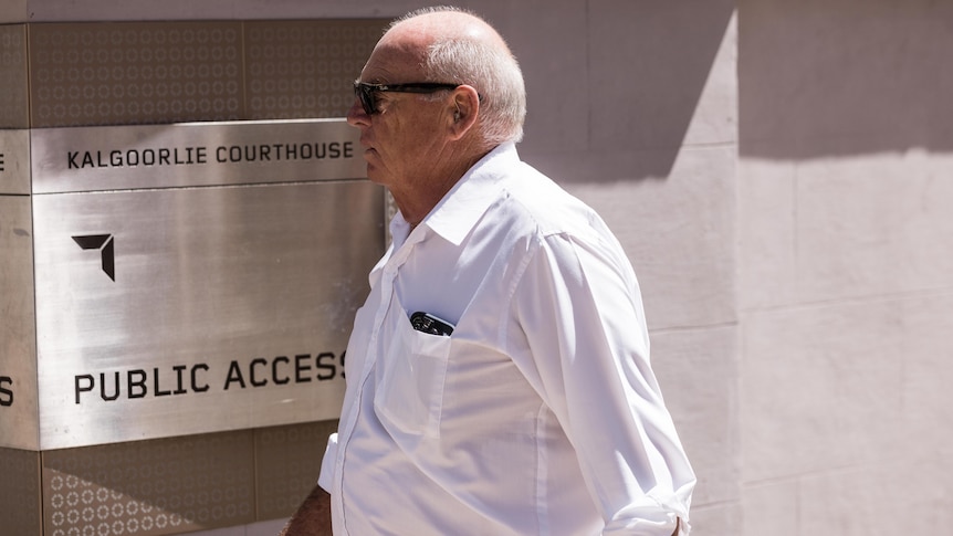 An older man wearing dark sunglasses and a business shirt walks away from a court building.