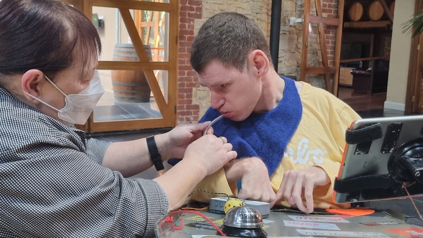 Man in a wheelchair sitting at a desk being assisted by a female support worker in a grey shirt to drink from a straw