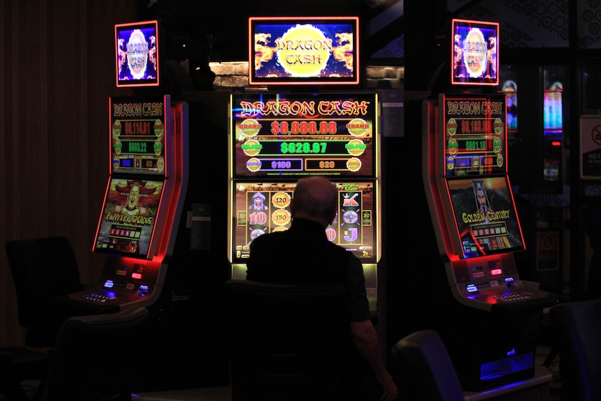 A man sits at a colourful poker machine. He is silhouetted by its light. Two other pokies sit next to it.