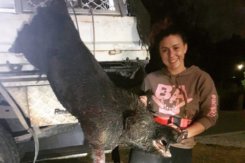 A women in her twenties holds a large pig that hangs upside down after being hunted. 