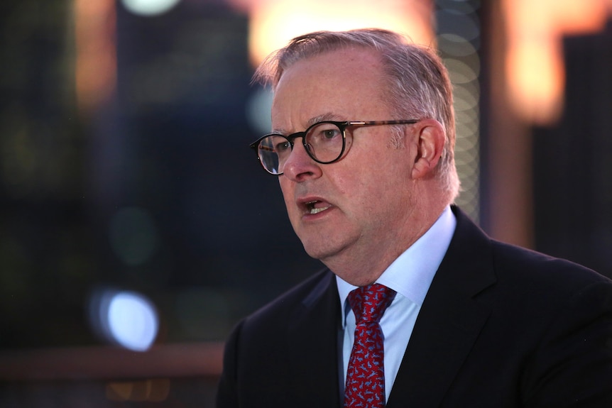 A middle-aged white man with grey hair and glasses wearing a suit speaks in a public place with a cityscape behind him.