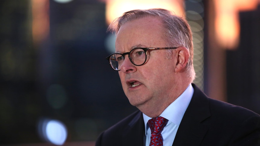 A middle-aged white man with grey hair and glasses wearing a suit speaks in a public place with a cityscape behind him.