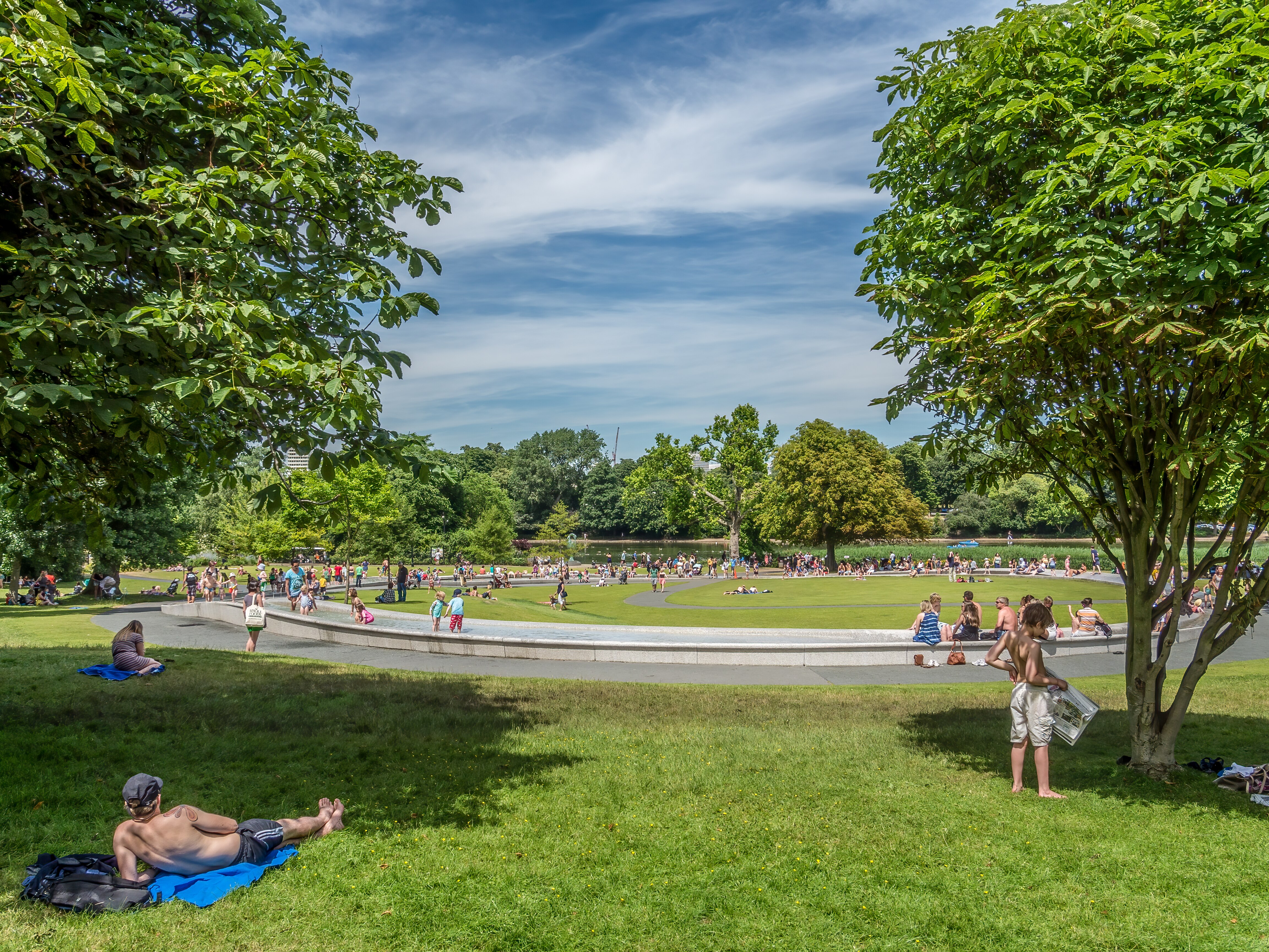 People lying on the grass near a round fountain under a sunny blue sky 