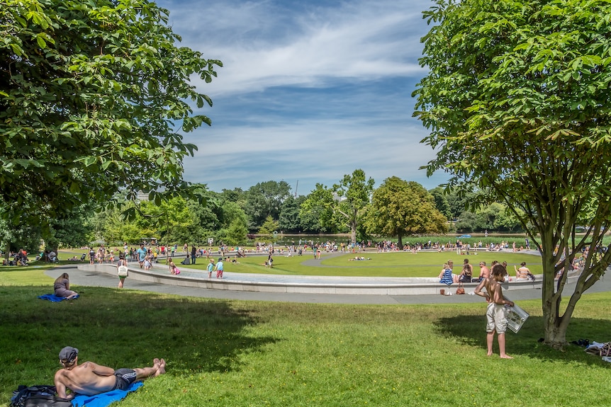 People lying on the grass near a round fountain under a sunny blue sky 