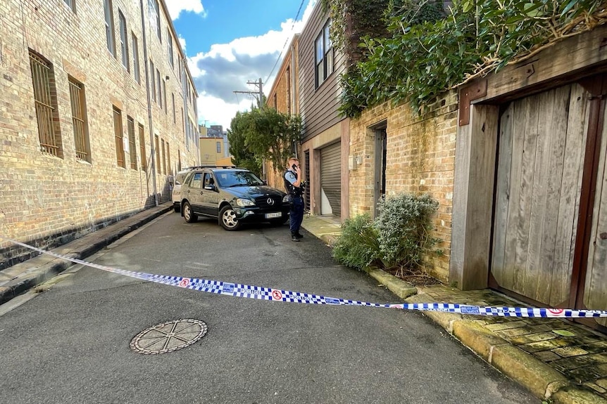 Crime scene with police tape in an alleyway with a black car and police officer in background