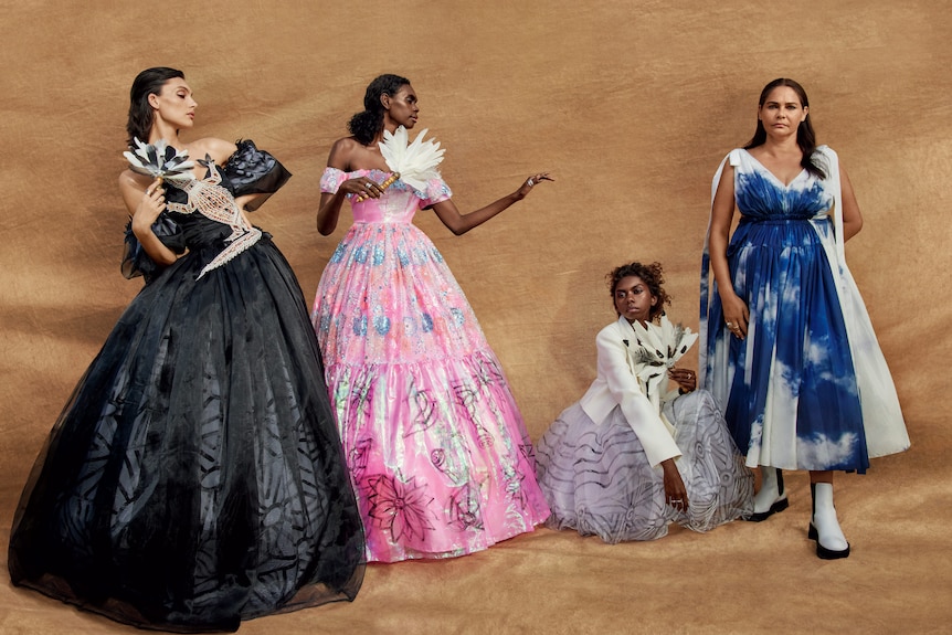 Four women in elaborate dresses pose in front of a beige background