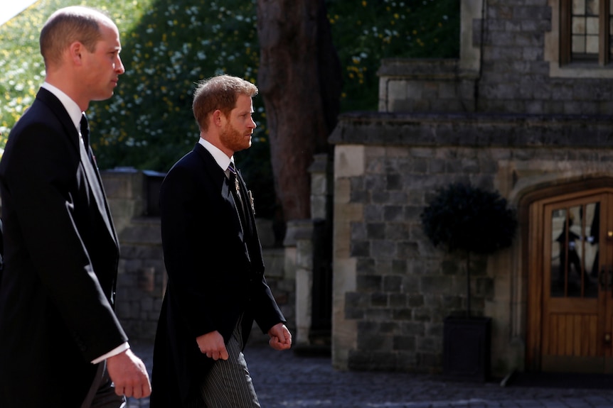 On a bright day, you view princes William and Harry in dark formal suits walking in front of stone buildings.