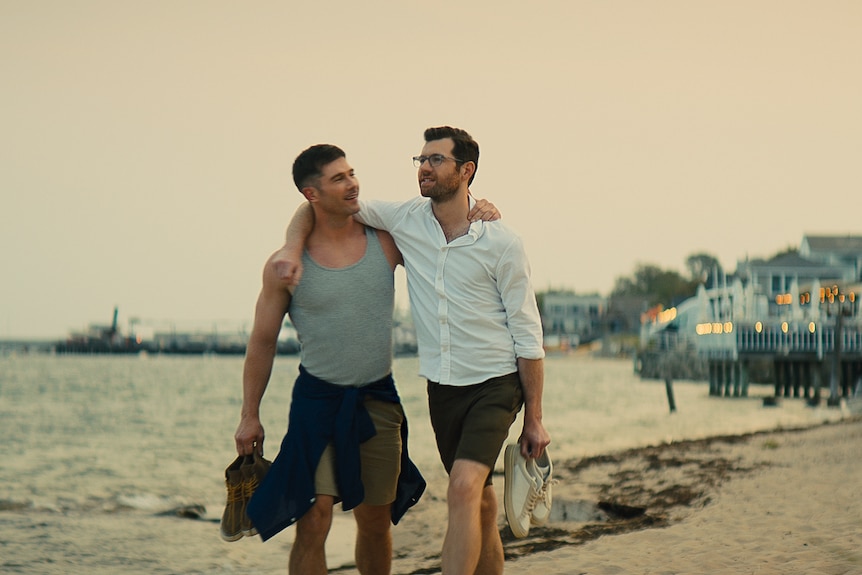 Two white men with short dark hair walk along a beach at dusk with their arms around each other's shoulders and holding shoes.
