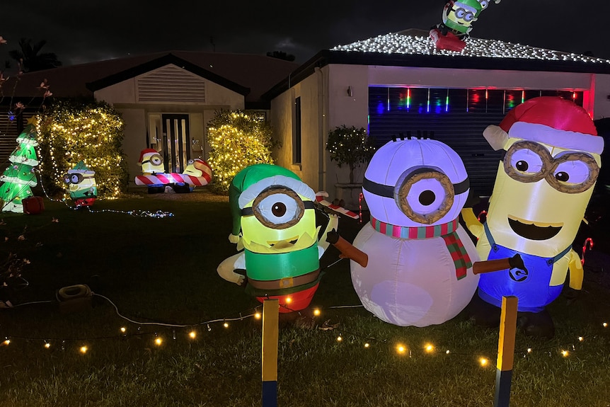 Blow up minion characters on a front lawn, houses lit up, decorated with Christmas lights behind.