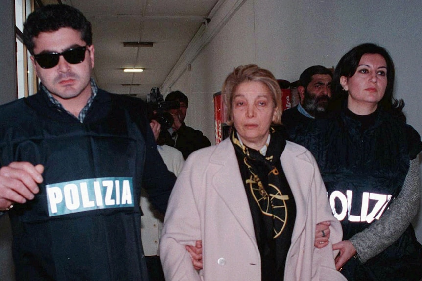 An older woman wearing a long pink cardigan and patterned scarf is escorted by officers wearing POLIZIA jackets