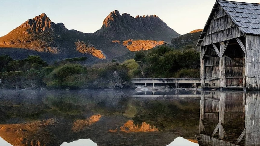 A wooden shed and surrounding trees, and a mountain in the background, are reflected in a lake.