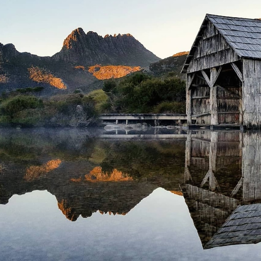 A wooden shed and surrounding trees, and a mountain in the background, are reflected in a lake.