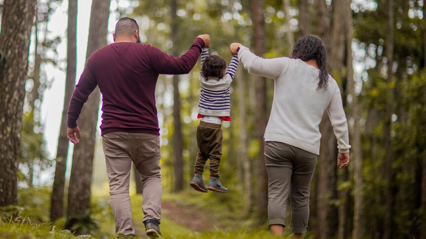 Two parents hold up a child by each hand as they walk through the woods.