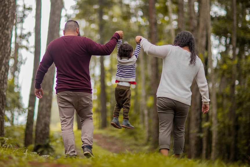 Two parents hold up a child by each hand as they walk through the woods.