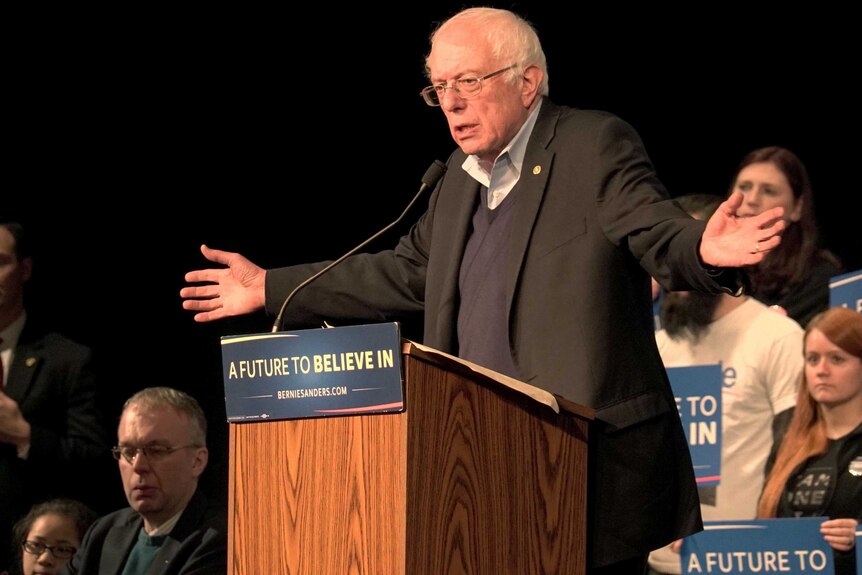 Bernie Sanders gestures as he speaks at a podium, with supporters behind him.