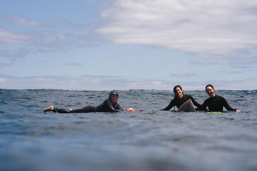 Three women wearing full length black wetsuits are lying on their boards in the ocean, looking at the camera smiling.