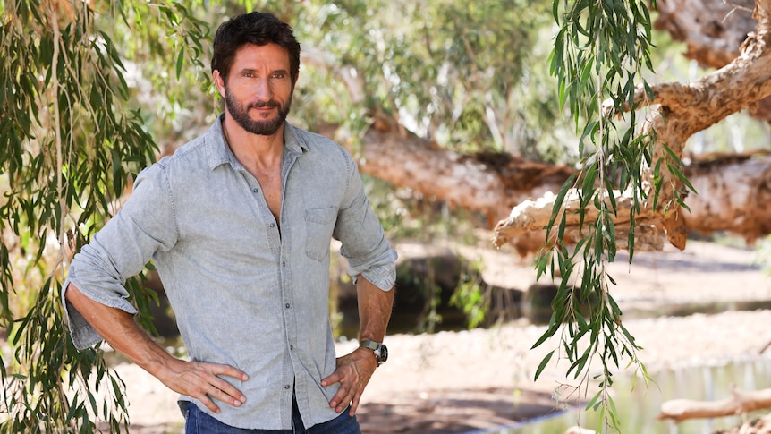 Australian Survivor host Jonathan LaPaglia stands with his hands on his hips next to a creek and paperbark tree
