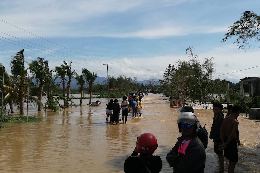 On a clear day, road is submerged by bright brown floodwaters as onlookers wade through angle-deep.