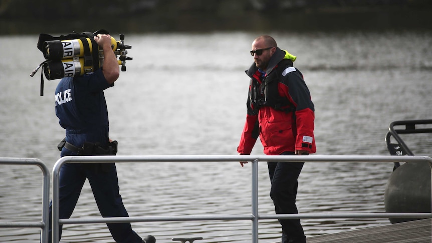 A police diver carrying oxygen tanks approaches a man wearing a red jacket on a jetty on the Swan River.
