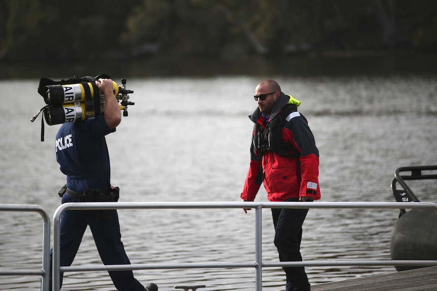 A police diver carrying oxygen tanks approaches a man wearing a red jacket on a jetty on the Swan River.