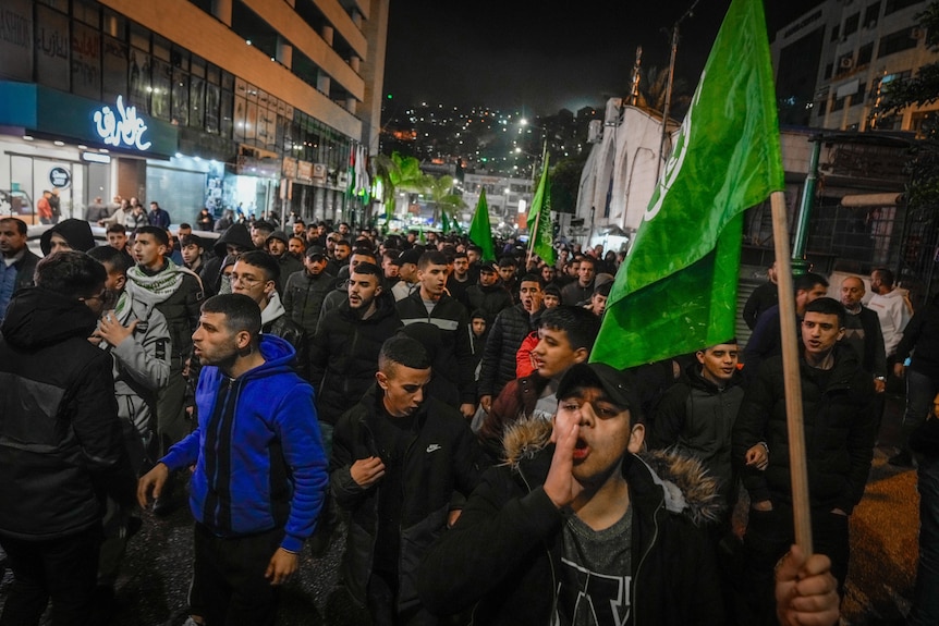 A group of men with green flags walk down a street at night 