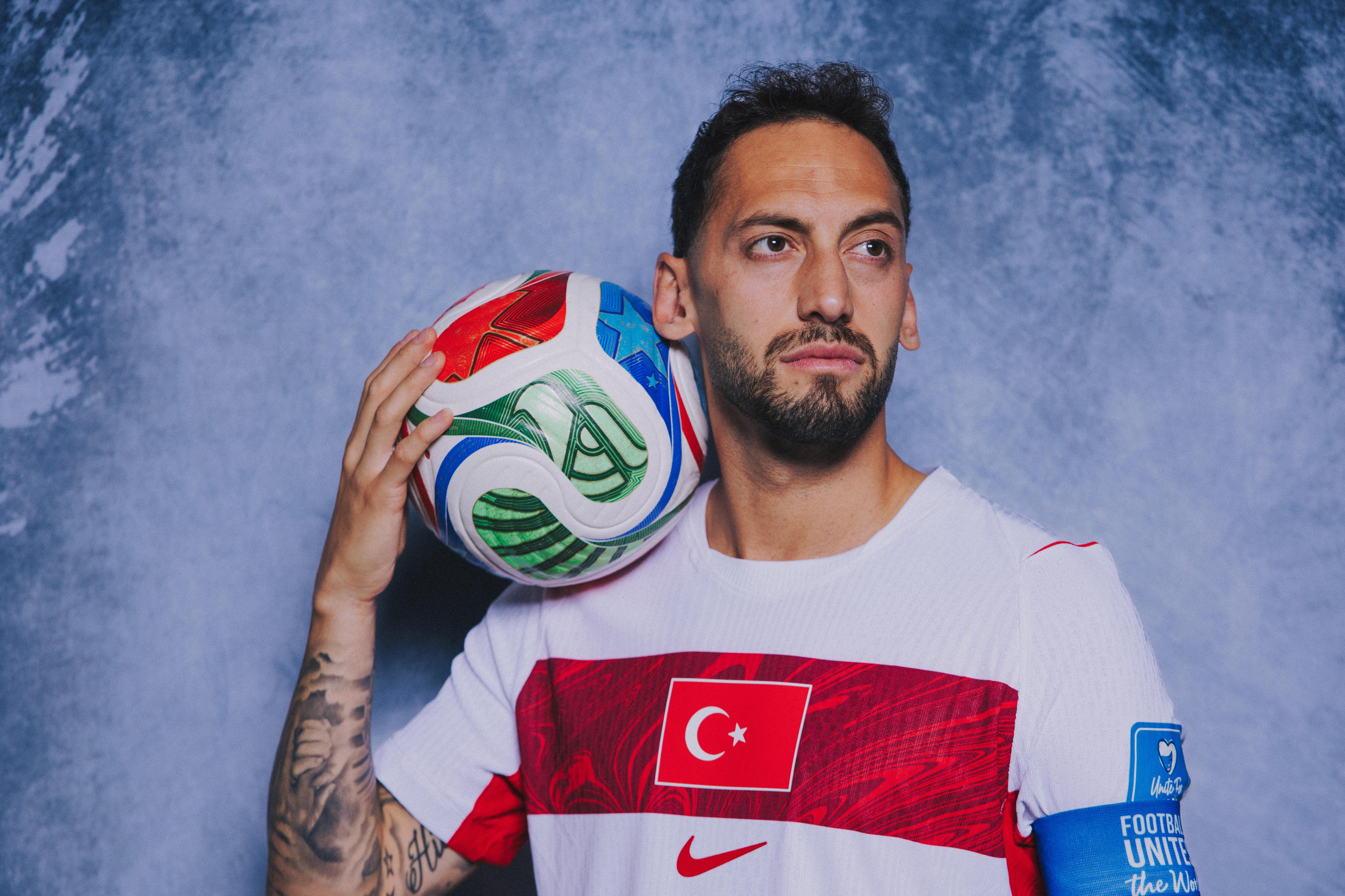 Footballer poses with the ball on his shoulder and neck, looking away from the camera.