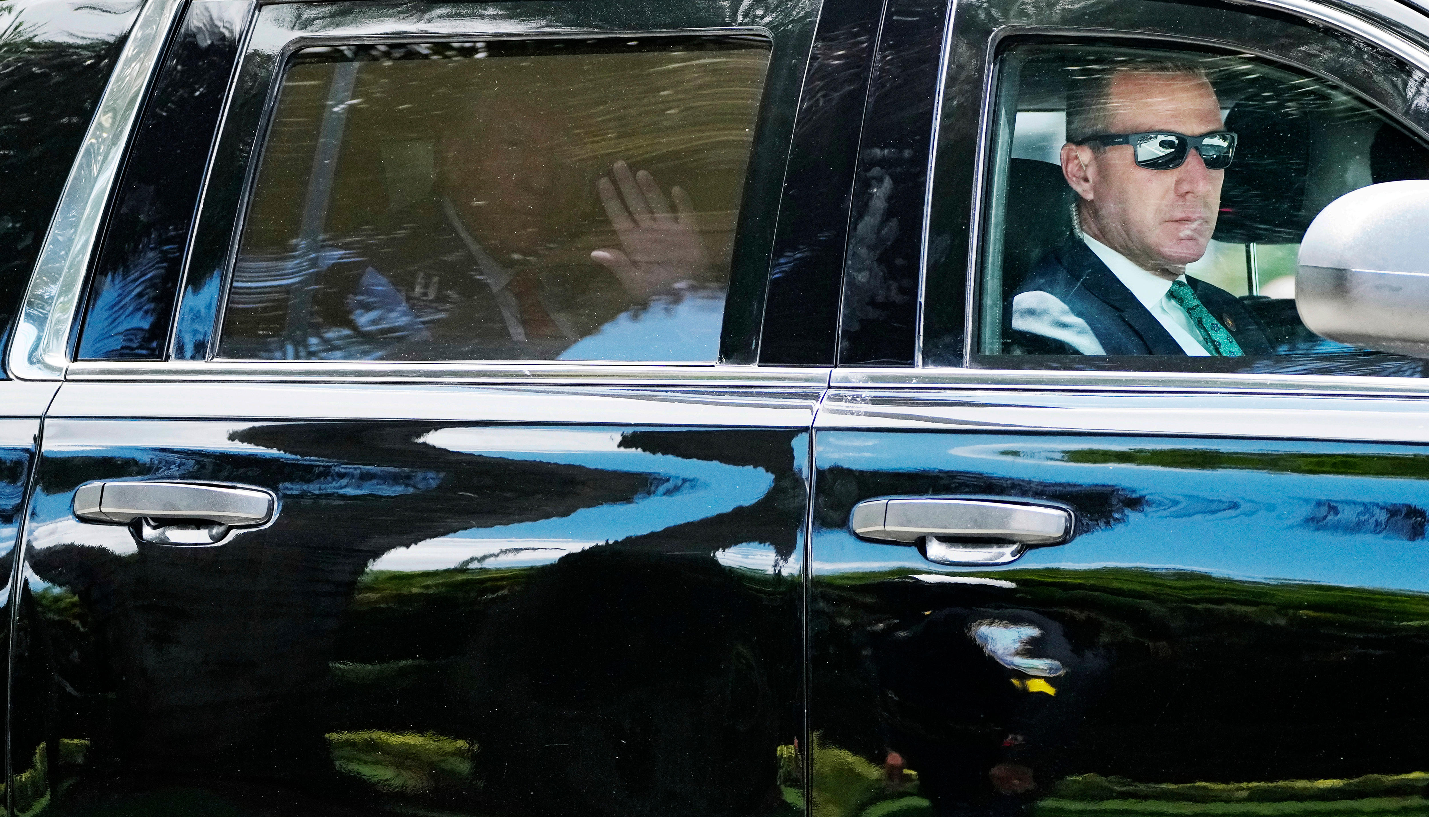 Donald Trump waves from the backseat of a limousine, while a driver with sunnies drives in the front seat.