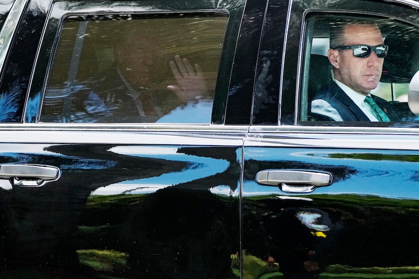 Donald Trump waves from the backseat of a limousine, while a driver with sunnies drives in the front seat.