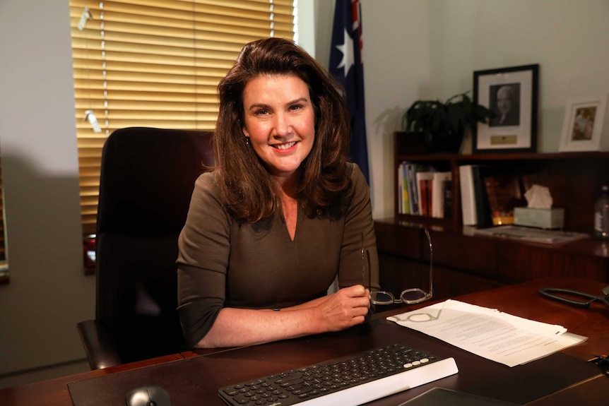 Jane Hume sits at her desk. One hand is holding reading glasses and the other is folded across her chest. She is smiling.