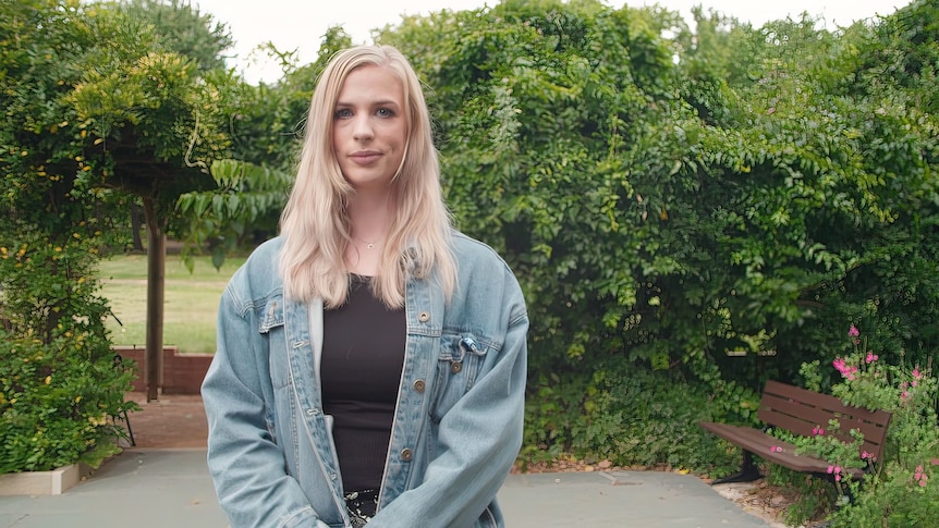 A woman in a denim jacket with blonde hair smiles stands outdoors.