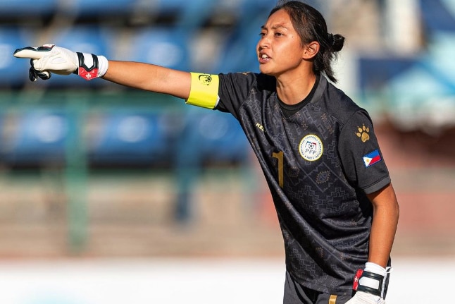 A female football goalkeeper stands and points, yelling instructions.