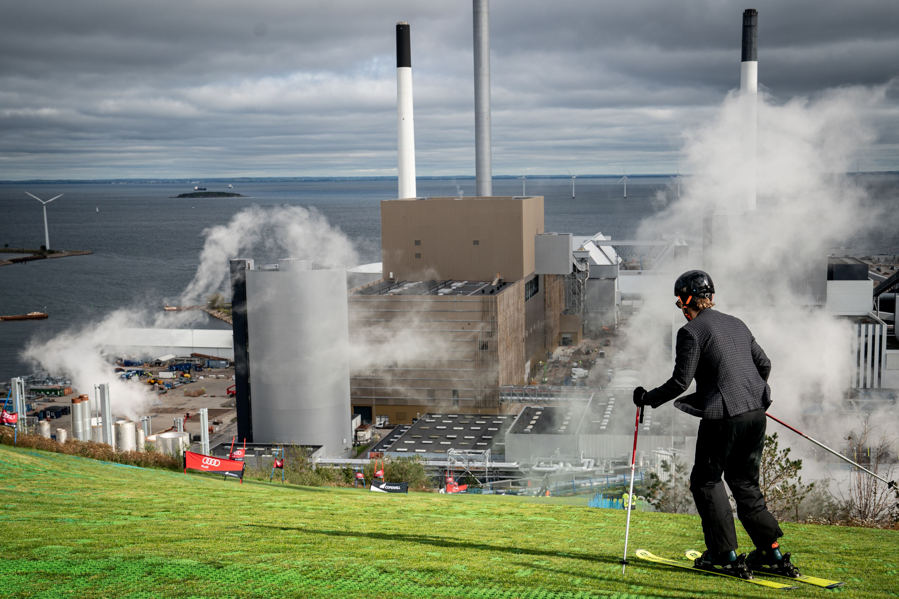 Un esquiador desciende una pendiente cubierta de hierba cerca de un sitio industrial que libera humo.
