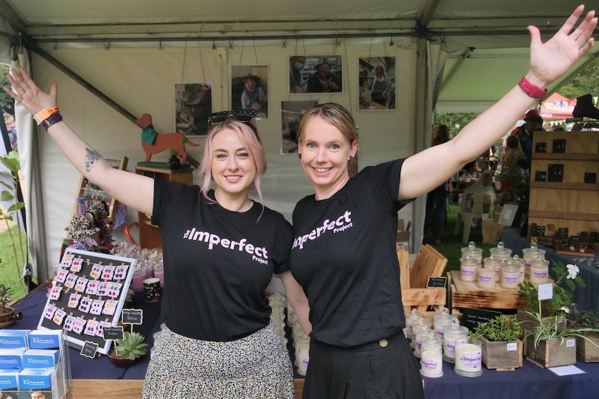 Two women with an arm raised in the air standing in front of a stall at a festival