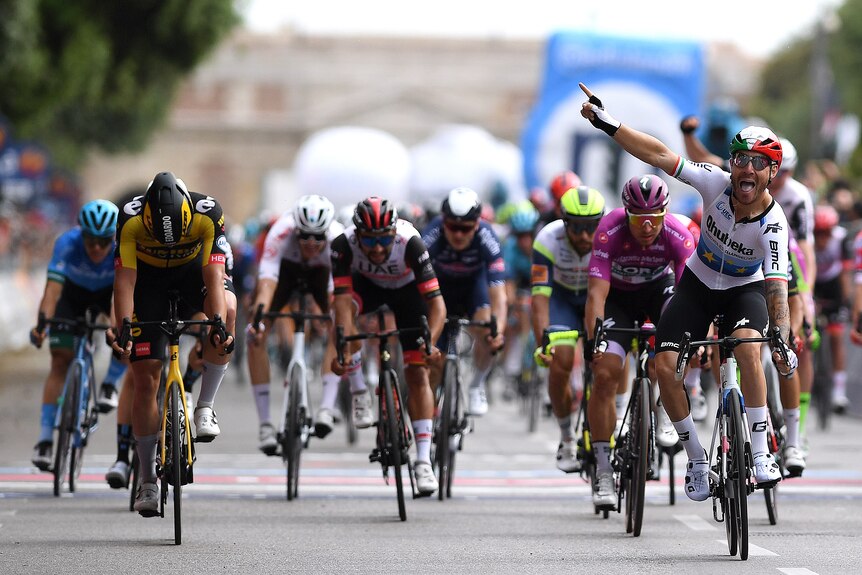 Giacomo Nizzolo levanta la mano con la boca abierta mientras otros ciclistas van detrás de él.