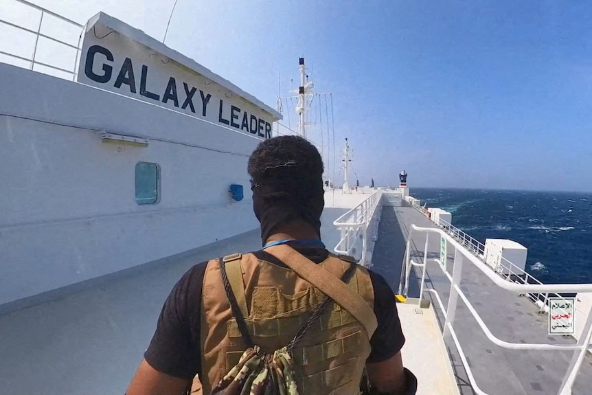 A Houthi rebel fighter standing with his back to the camera on the deck of a cargo ship. 